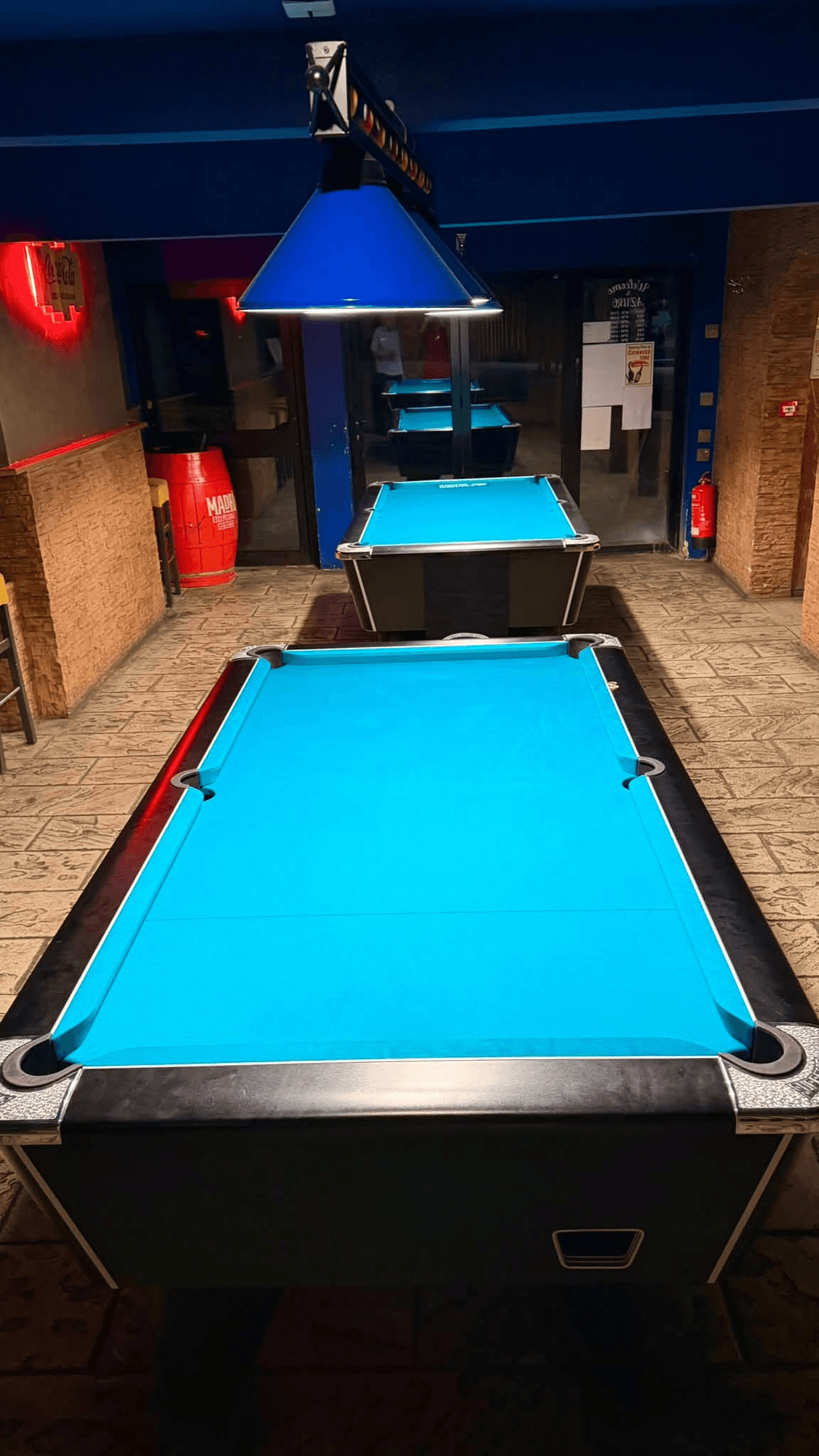 Two pool tables with blue cloth under a blue lamp